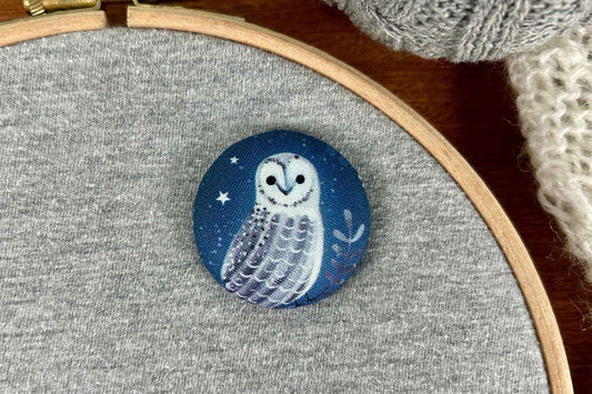 A round fabric magnetic needle minder featuring a white owl under a dark blue night sky, displayed on a gray fabric embroidery hoop.