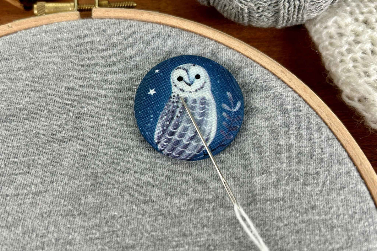 A close-up of a circular fabric magnetic needle minder featuring a white owl on a dark blue night sky, pinned onto a gray knit garment inside an embroidery hoop with a needle visible.