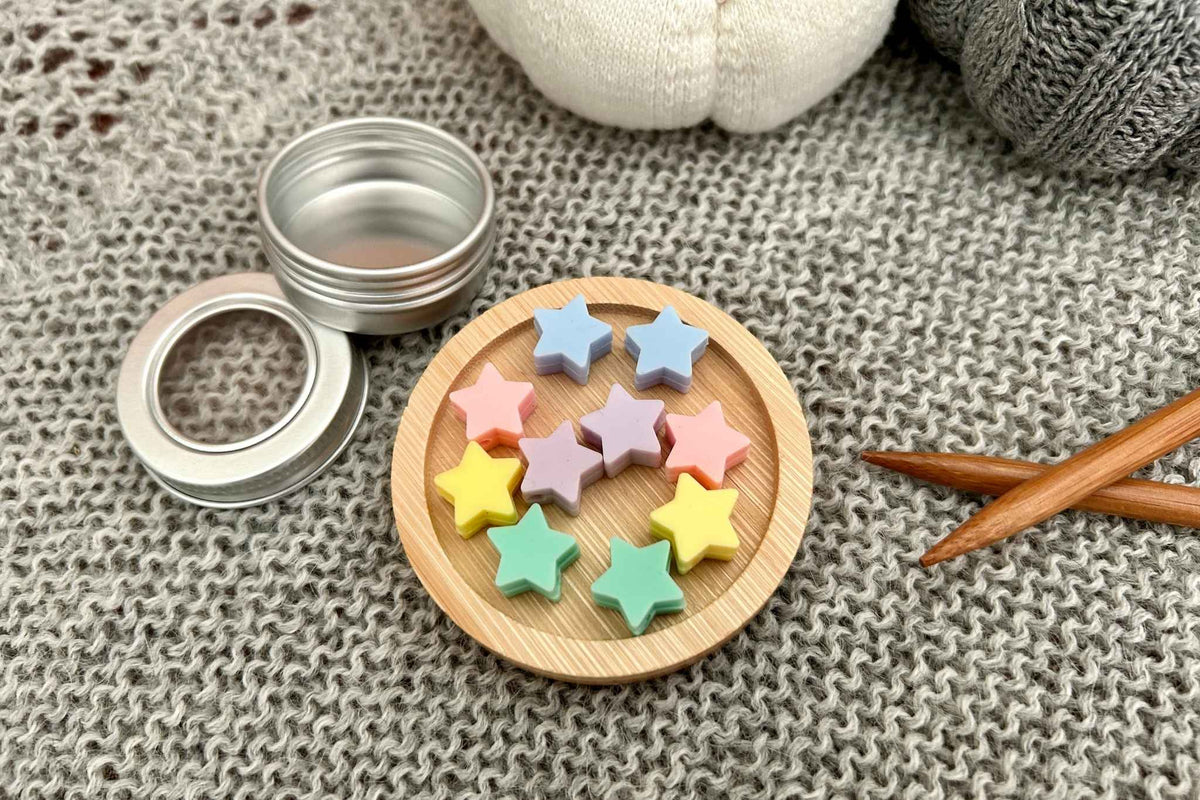 A set of 10 pastel star-shaped knitting needle stoppers arranged on a small round wooden dish, with an open silver metal tin and its lid beside it on a gray knitted blanket. Wooden knitting needles and a white knitted pumpkin are also visible in the background.