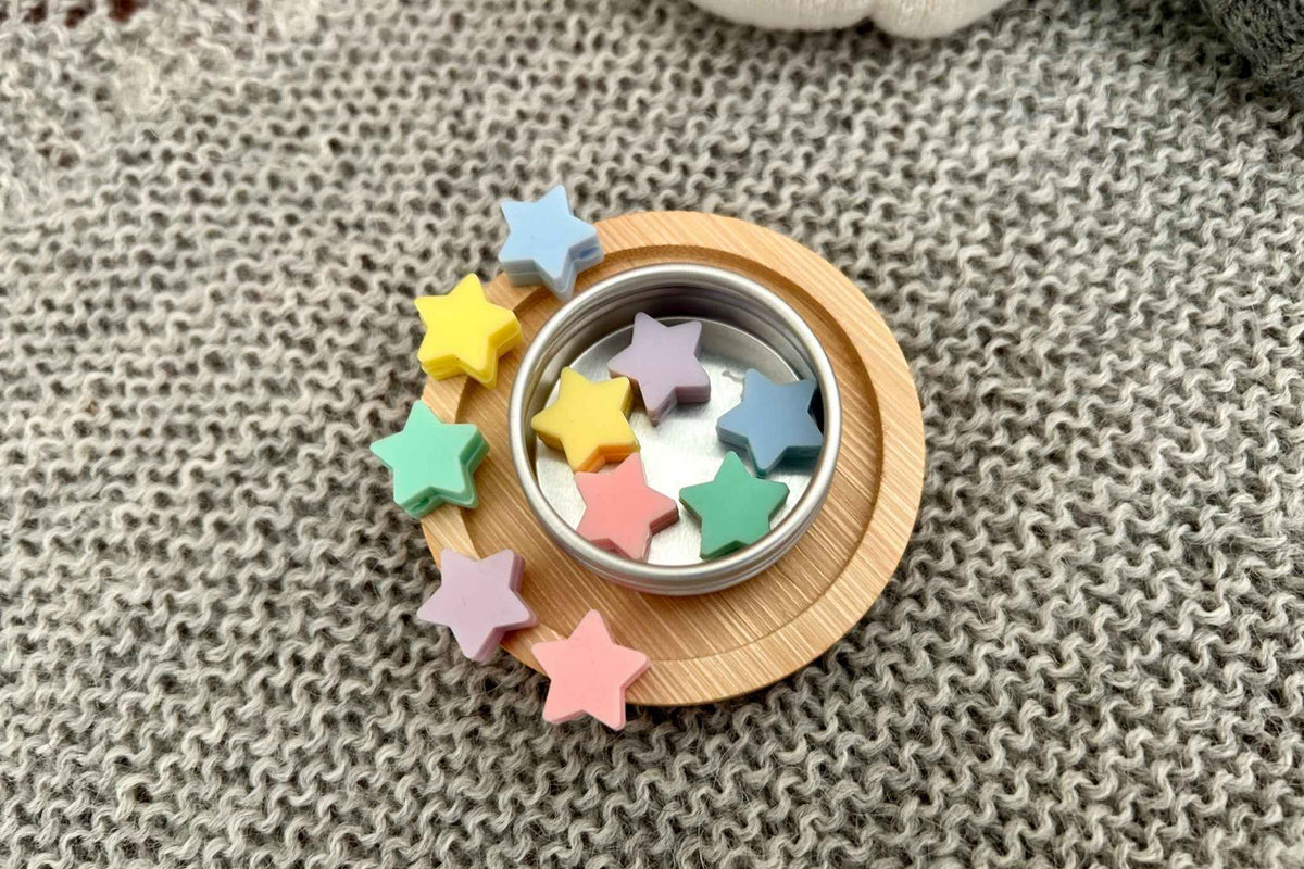 A small round metal tin with 10 pastel star-shaped knitting needle stitch savers inside, placed on a round wooden coaster over a gray knitted fabric background.