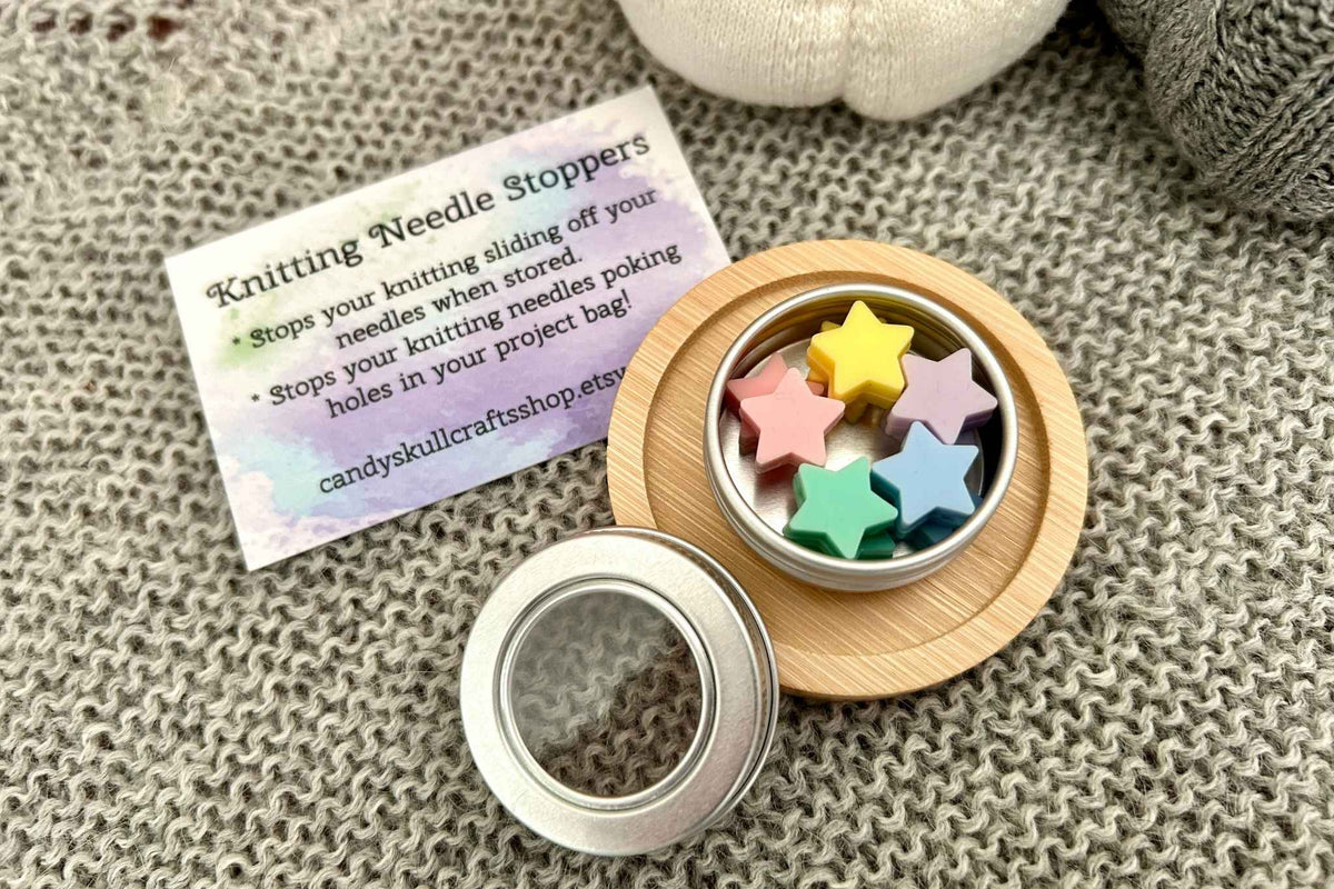 A small round metal tin with 10 knitting needle stoppers sits on a wooden coaster beside a printed product card on a knitted gray surface. The open lid is placed in front, and the tin contains pink, yellow, lilac, mint green, and blue stoppers. 