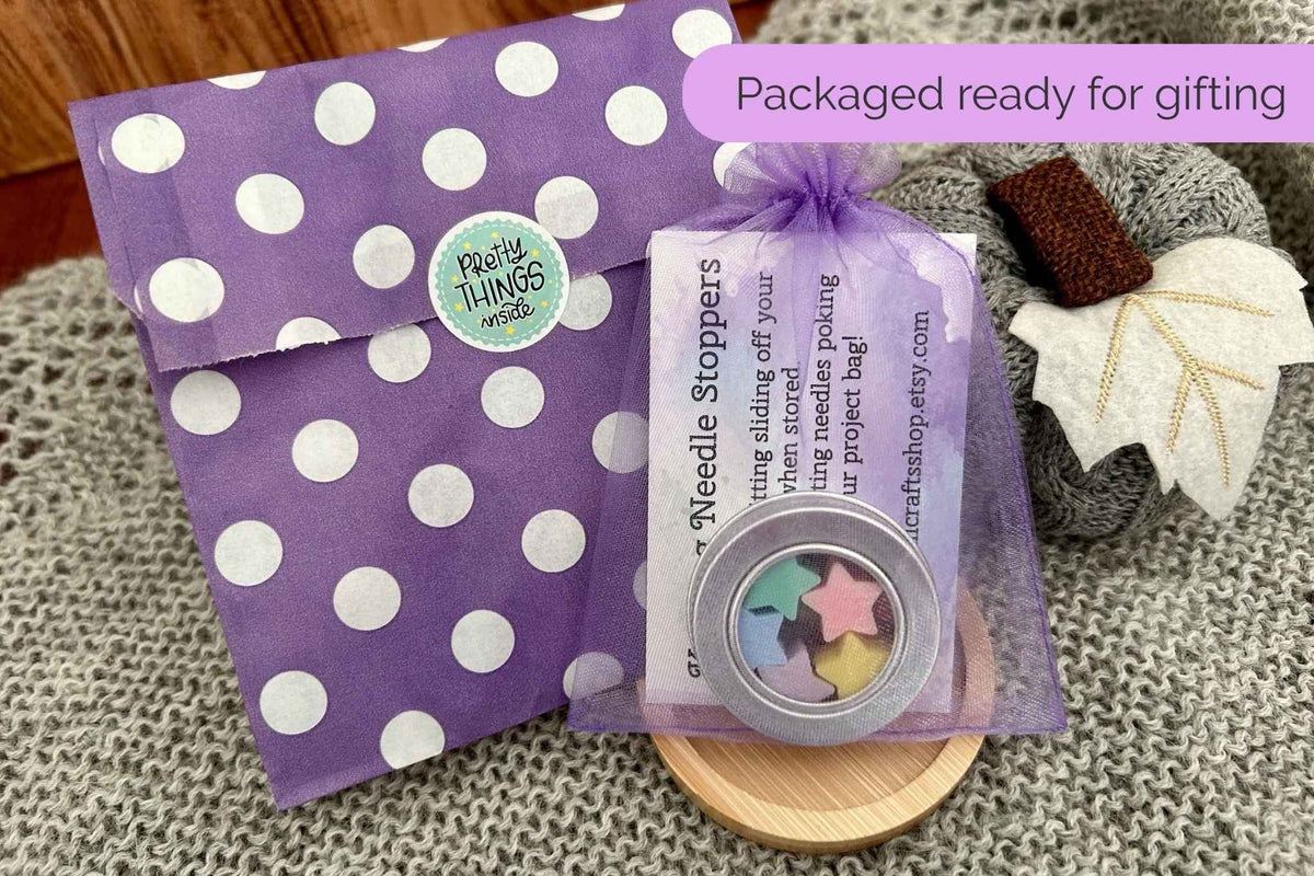 Purple polka dot paper bag and a sheer organza bag holding a round needle stopper tin, styled on a gray knit blanket with a wooden surface and decorative leaf props. Text reads "packaged ready for gifting"