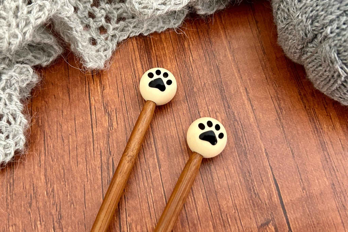 Two wooden knitting needles with round cream-colored paw print end caps or knitting needle stoppers, displayed on a brown wooden surface with gray knitted yarn in the background.