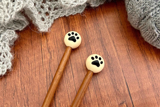 Two wooden knitting needles with round cream-colored paw print end caps or knitting needle stoppers, displayed on a brown wooden surface with gray knitted yarn in the background.