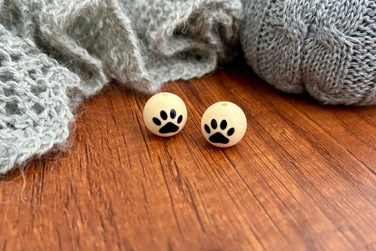 Two round cream-colored knitting needle stitch savers with black paw prints, displayed on a wooden surface next to gray knitted blankets.