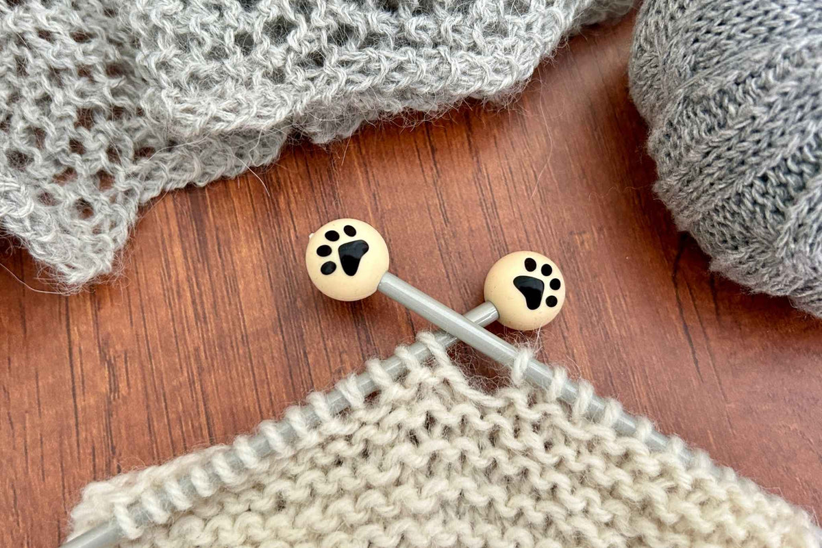 Close-up of knitting needles with two round stitch savers featuring black paw print designs stopping the knitting sliding off the needles, resting on a wooden surface with knitted fabric and yarn nearby.