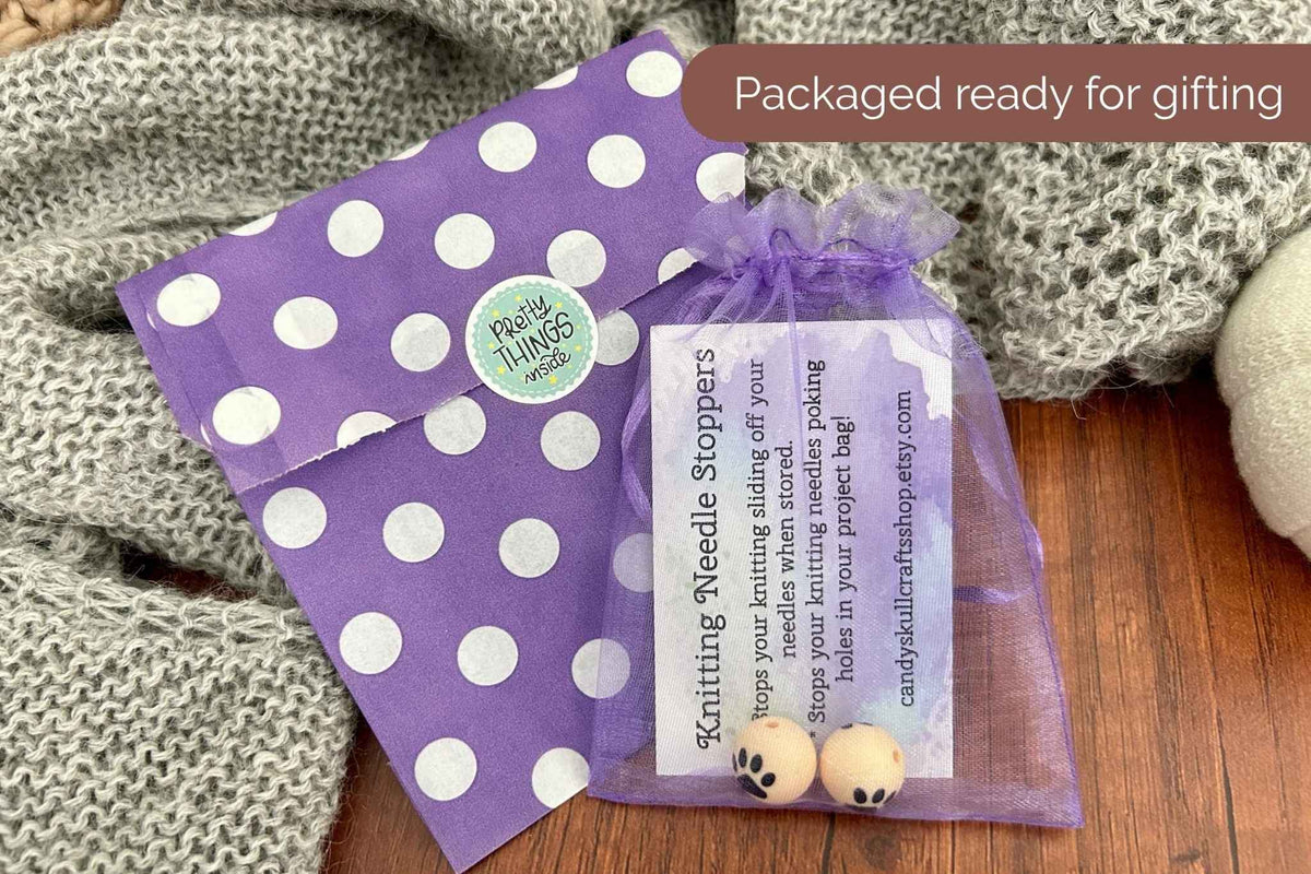 Purple and white polka dot gift packaging with a small purple organza pouch containing a printed card and two paw print knitting needle stoppers, styled on a wooden surface with a gray knit blanket.