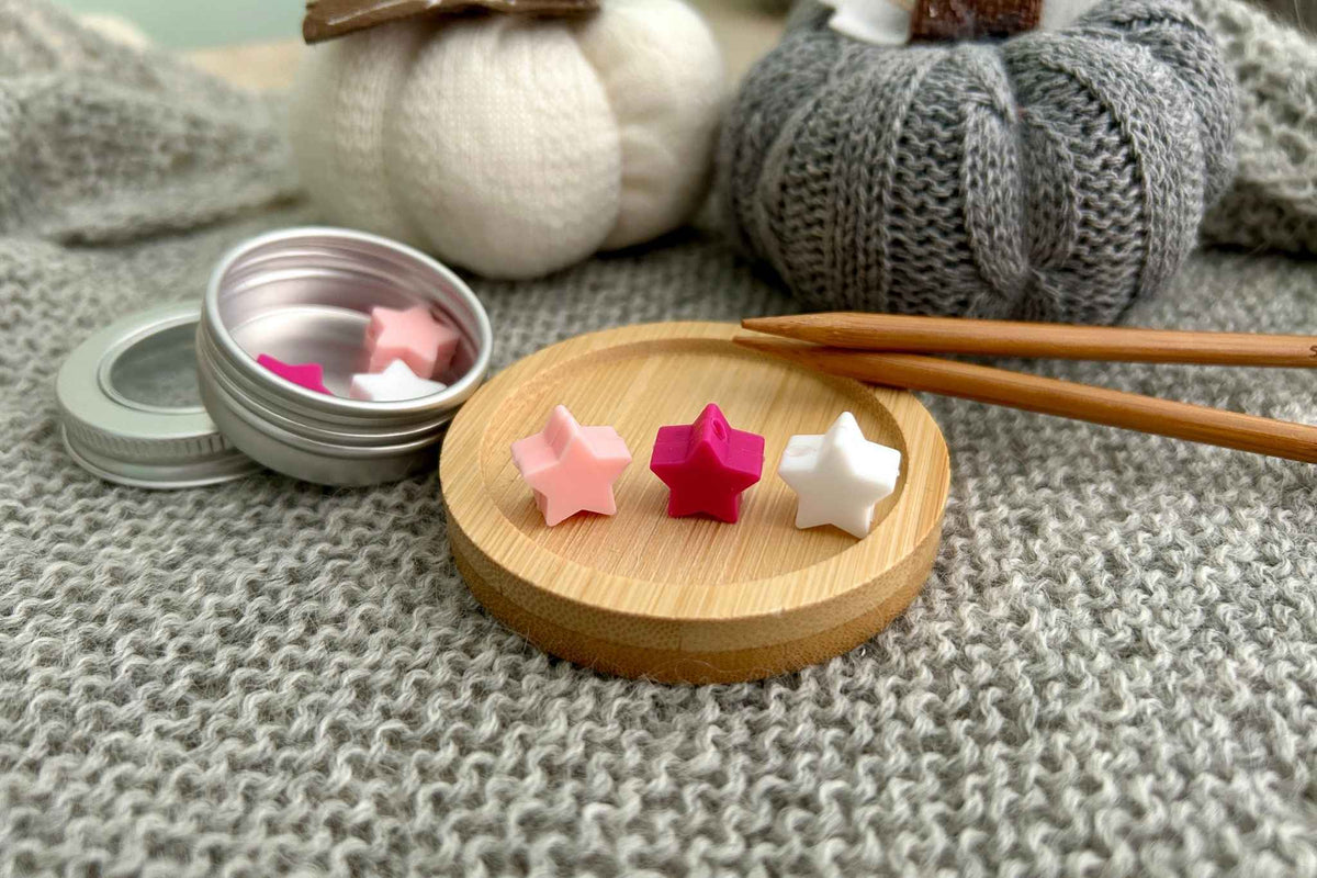 Three small star-shaped knitting needle stoppers in pink, magenta, and white are displayed on a round wooden tray, with a metal tin of the toys and wooden knitting needles nearby on a gray knitted blanket. Decorative knitted pumpkins sit in the background.