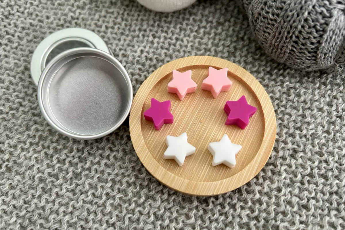Four small star-shaped knitting needle stoppers in pink, magenta, and white, arranged on a round bamboo tray next to an open silver metal tin, set on a gray knit blanket.