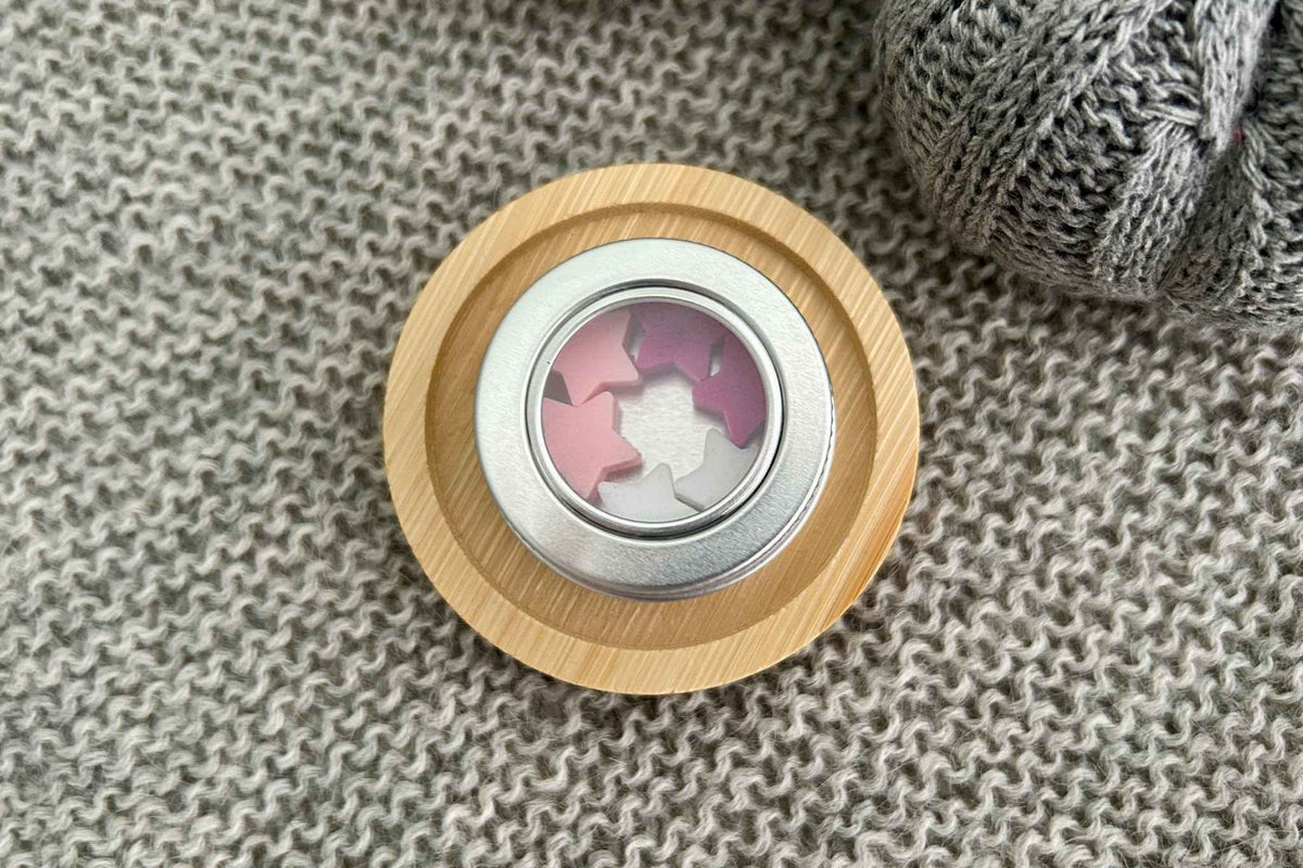 A circular metal tin with a window in the lid showing 6 pink star knitting needle stoppers, placed on a gray knitted fabric next to a gray knit item.