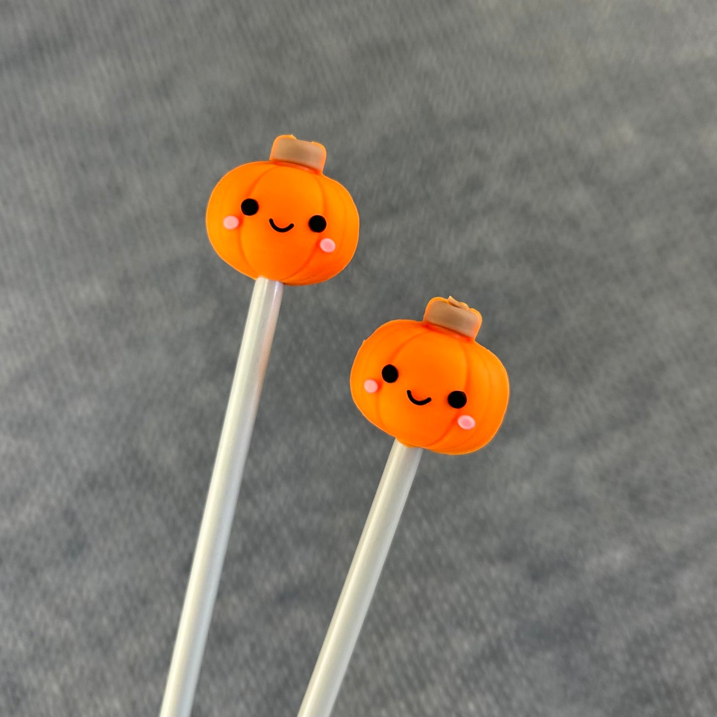 Two silicone knitting needle stoppers shaped like pumpkins with smiley faces and pink cheeks on the top, attached to the ends of metal knitting needles.
