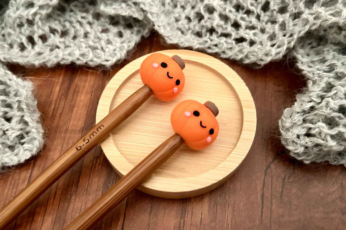 Two wooden knitting needles with orange pumpkin-shaped stitch savers with smiling faces, resting on a small round wooden plate over a dark wood surface with gray knitted fabric in the background.