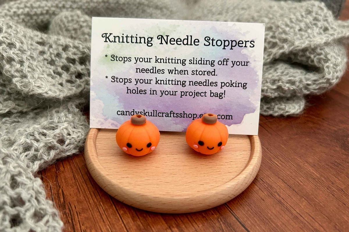 Two small orange pumpkin-shaped knitting needle stoppers with cute faces sit on a round wooden stand in front of a product card that reads 'Knitting Needle Stoppers.' The setup is styled on a wooden surface with a gray knitted fabric in the background.