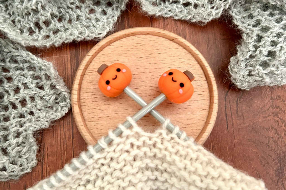 Two small orange knitted pumpkin-shaped knitting needle stoppers or savers with cute faces resting on crossed knitting needles over a wooden coaster, with knitted fabric in the foreground and gray knitted pieces around them.