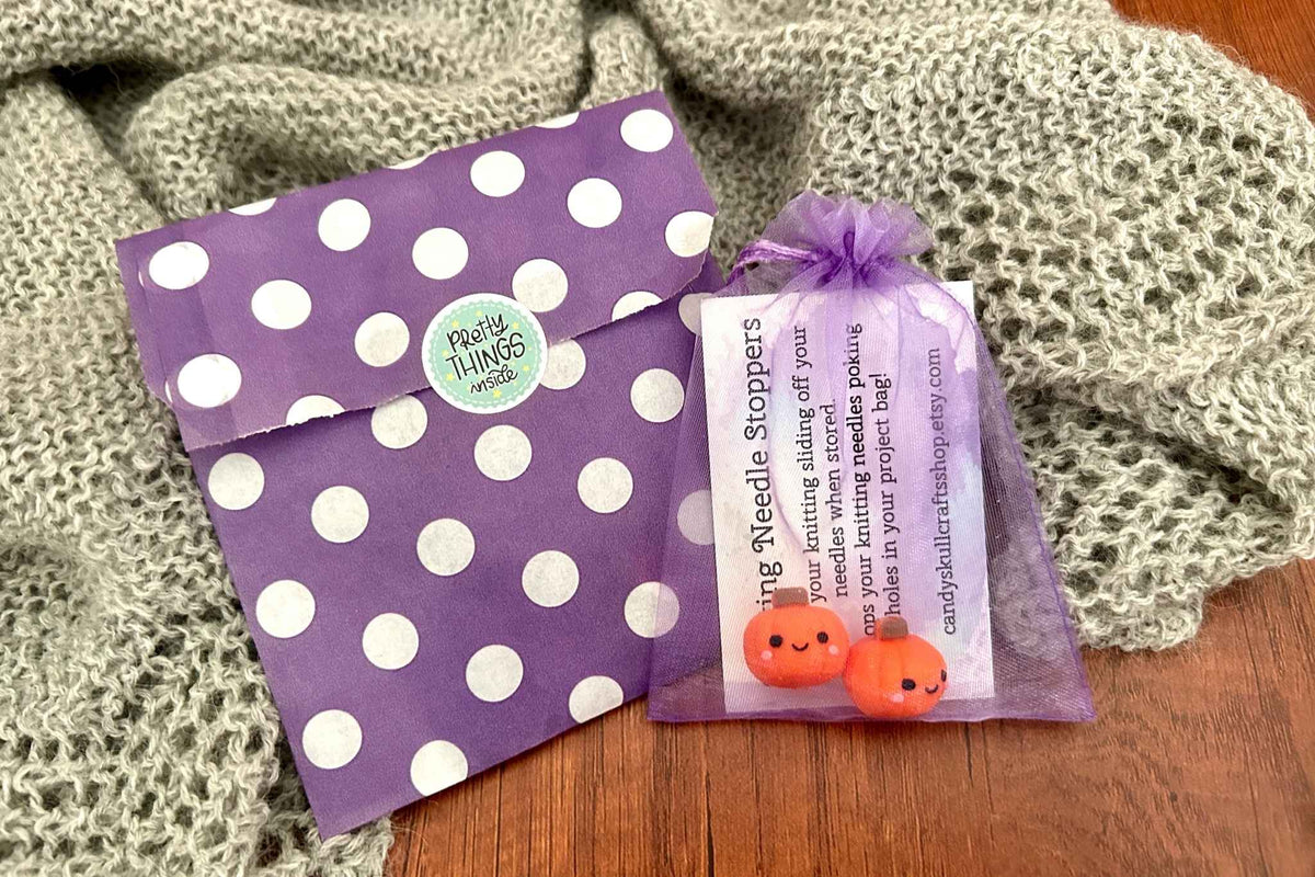 A purple and white polka dot favour bag placed next to a clear purple organza bag containing orange pumpkin-shaped needle stoppers and a product info card, set on a wooden table with a knitted blanket in the background.