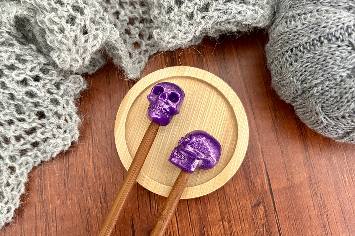 Two wooden knitting needles with metallic purple skull-shaped needle toppers or stitch savers resting on a round wooden coaster, surrounded by gray knitted yarn on a dark wood surface.