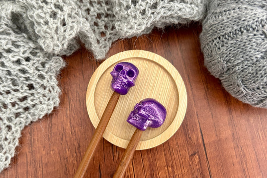 Two wooden knitting needles with metallic purple skull-shaped needle toppers or stitch savers resting on a round wooden coaster, surrounded by gray knitted yarn on a dark wood surface.