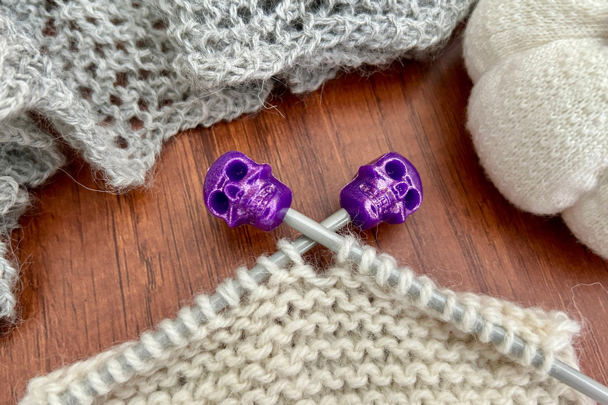 Close-up of knitting needles with shiny purple skull-shaped needle caps, working through cream-colored yarn on a wooden surface, with gray knitted fabric and a pumpkin in the background.