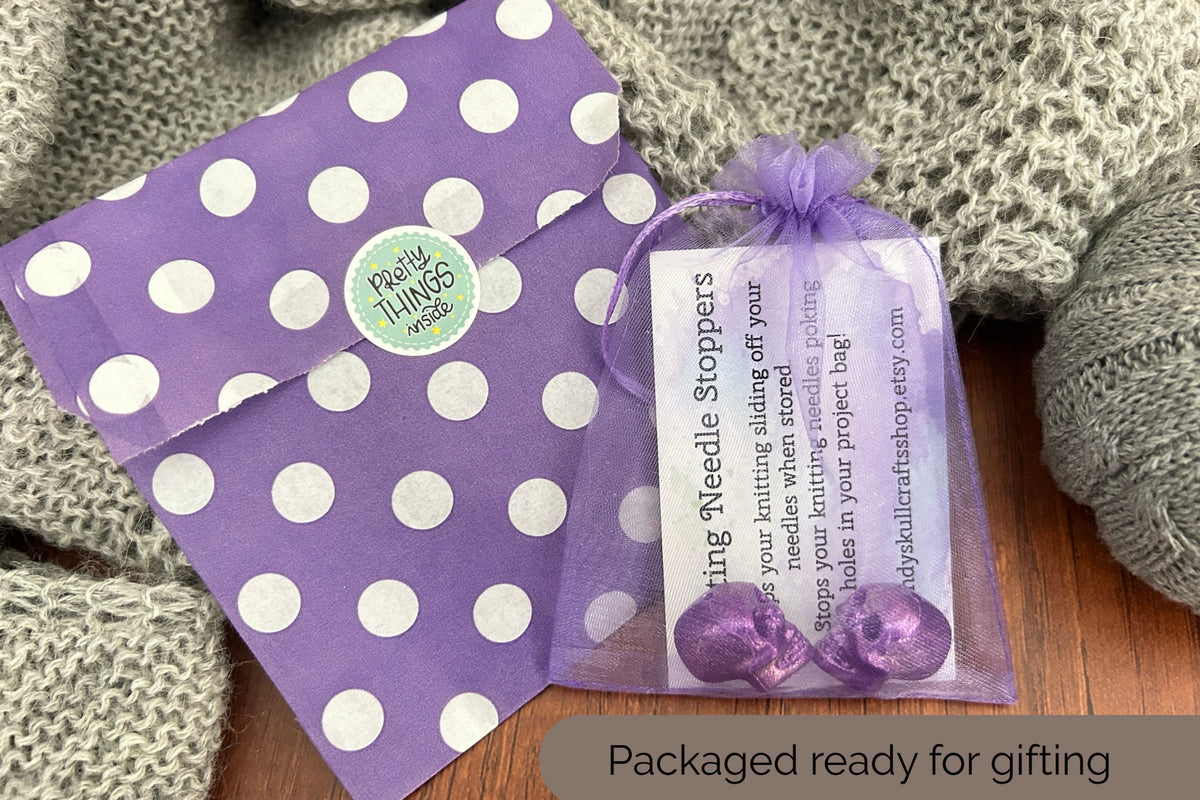 Purple and white polka dot gift packaging with a small tulle bag containing two purple skull shaped  needle stoppers and a product tag, styled on a wooden surface with a gray knitted blanket.
