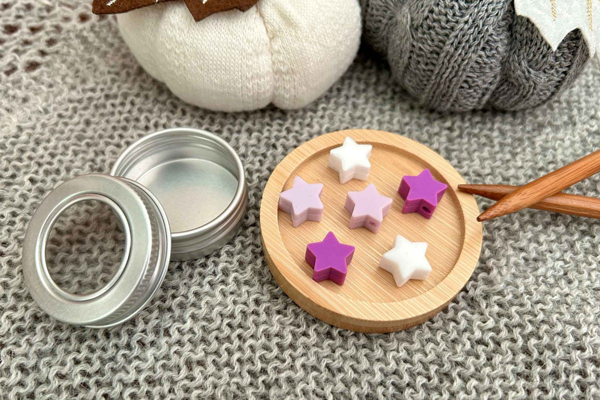A set of 6 star-shaped knitting needle stoppers in white, lilac and purple, shown with a small silver metal tin and a wooden display tray on a gray knitted blanket.