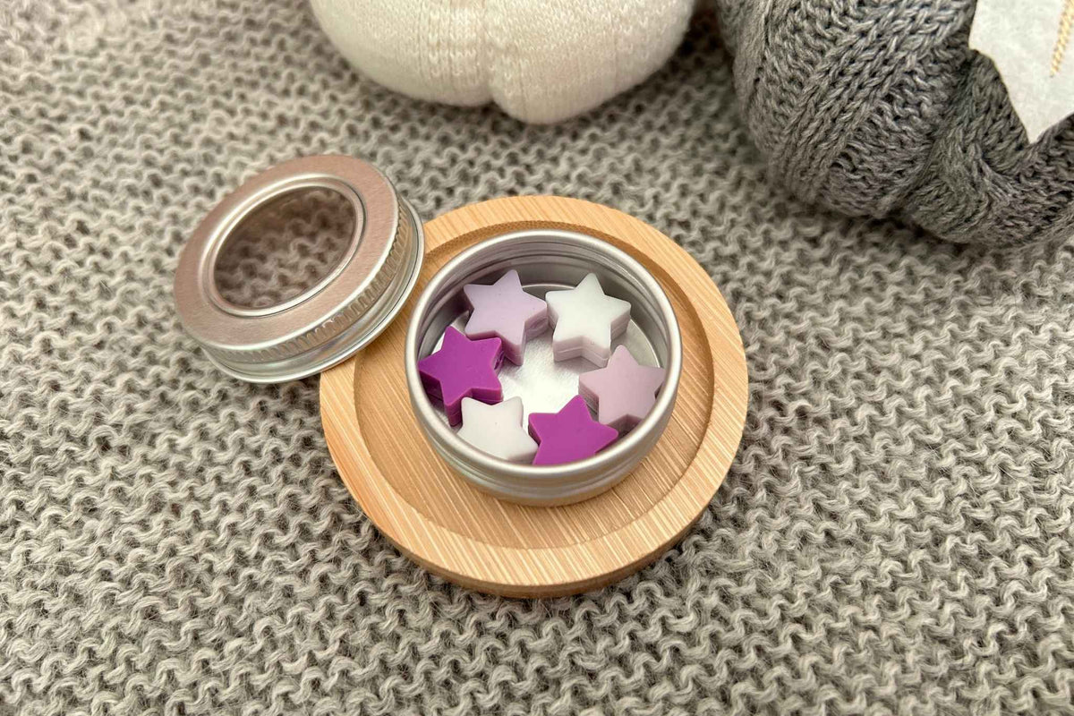 A small round metal tin with a wooden base holding 6 star-shaped knitting stitch savers in white, lilac, and purple, shown on a gray knit blanket with the lid placed beside it.
