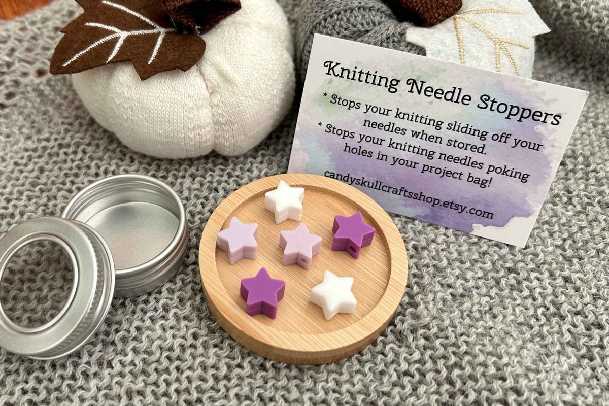 A set of six star-shaped knitting needle stoppers in white, lilac, and purple, displayed on a small wooden dish next to a metal tin lid and a product instruction card, arranged on a gray knit blanket.