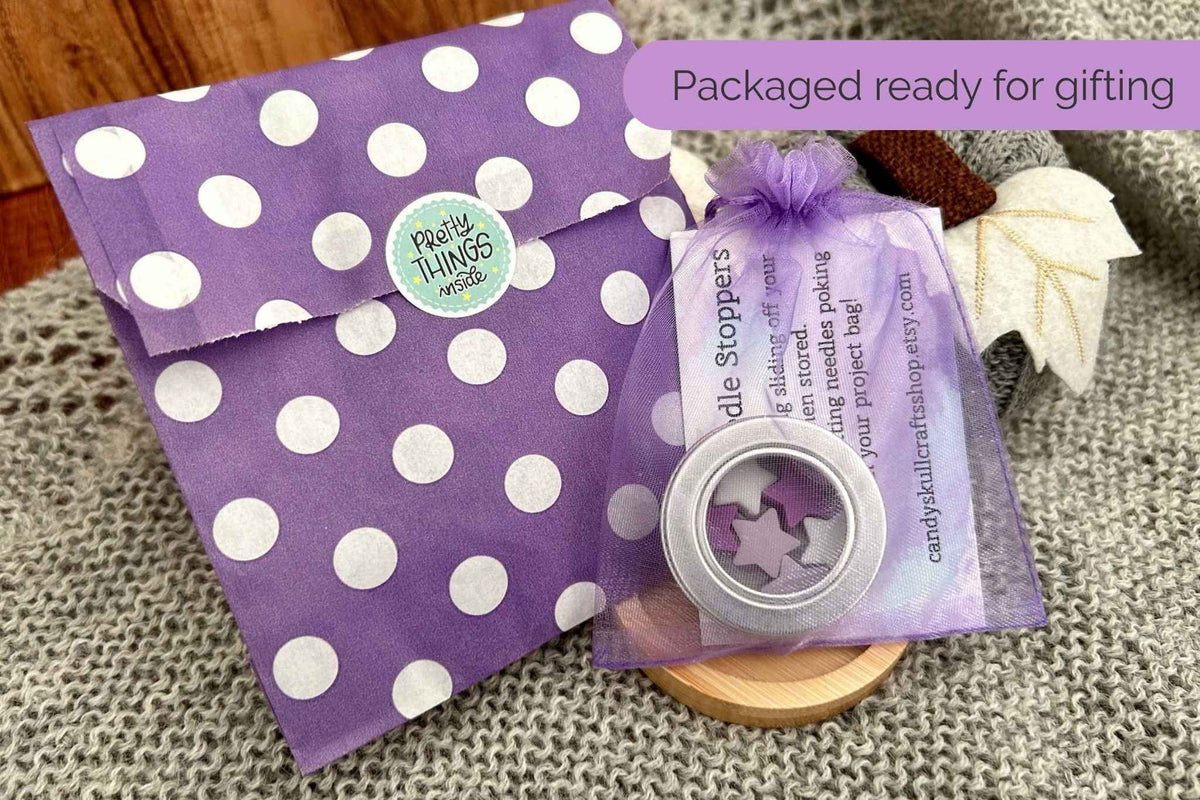 Purple and white polka dot gift packaging with a small organza bag containing a silver tin with purple, lilac, and white stitch savers, displayed on a textured fabric surface with a wooden background.