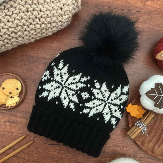 A black knit beanie with a large fluffy black pom-pom and a white Selbu star or snowflake pattern across the upper half, laid flat on a wooden surface with cosy props around it. The pattern takes the Selbu motif from Norwegian stranded colourwork traditions