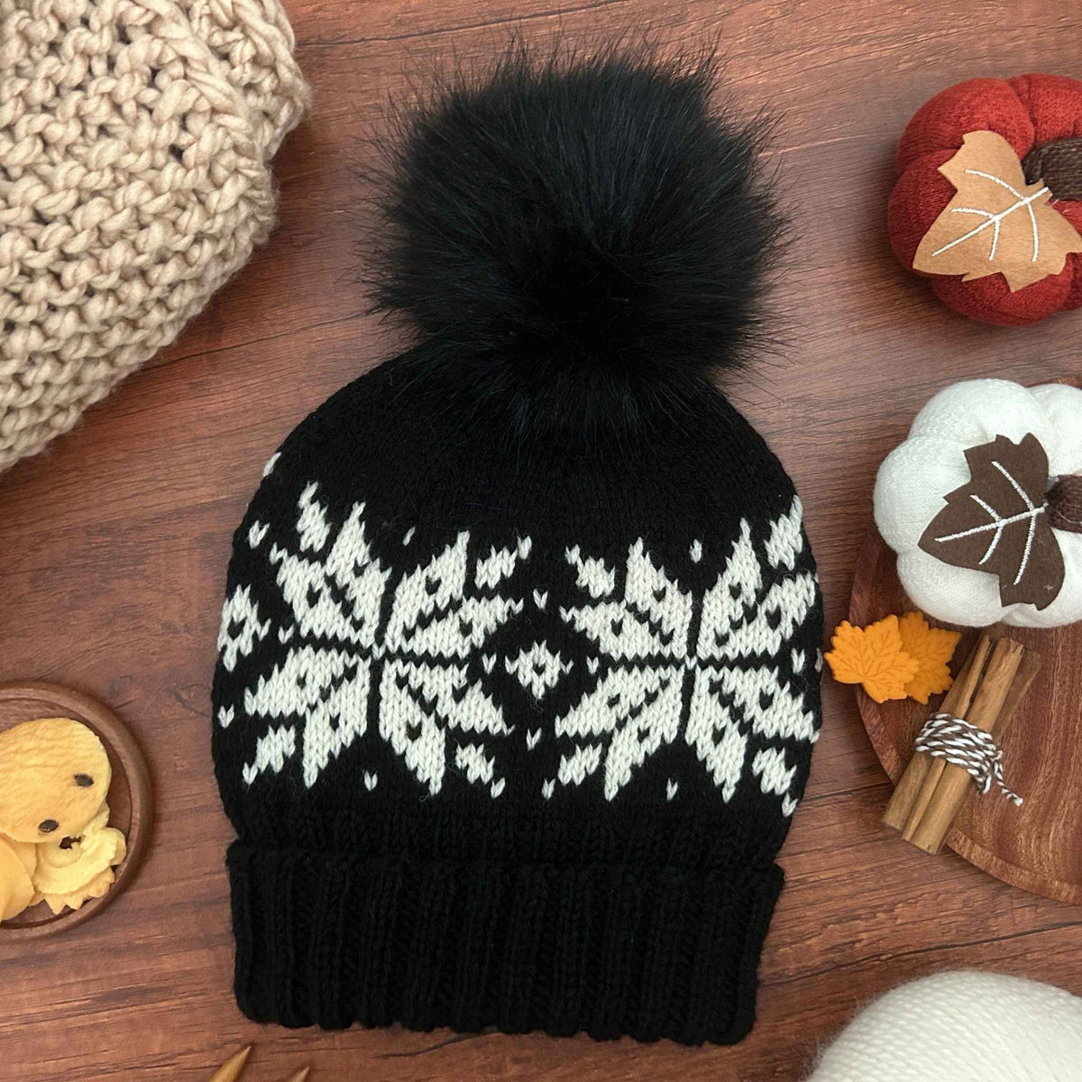 A black knit beanie with a large fluffy black pom-pom and a white Selbu star or snowflake pattern across the upper half, laid flat on a wooden surface with cosy props around it. The pattern takes the Selbu motif from Norwegian stranded colourwork traditions.