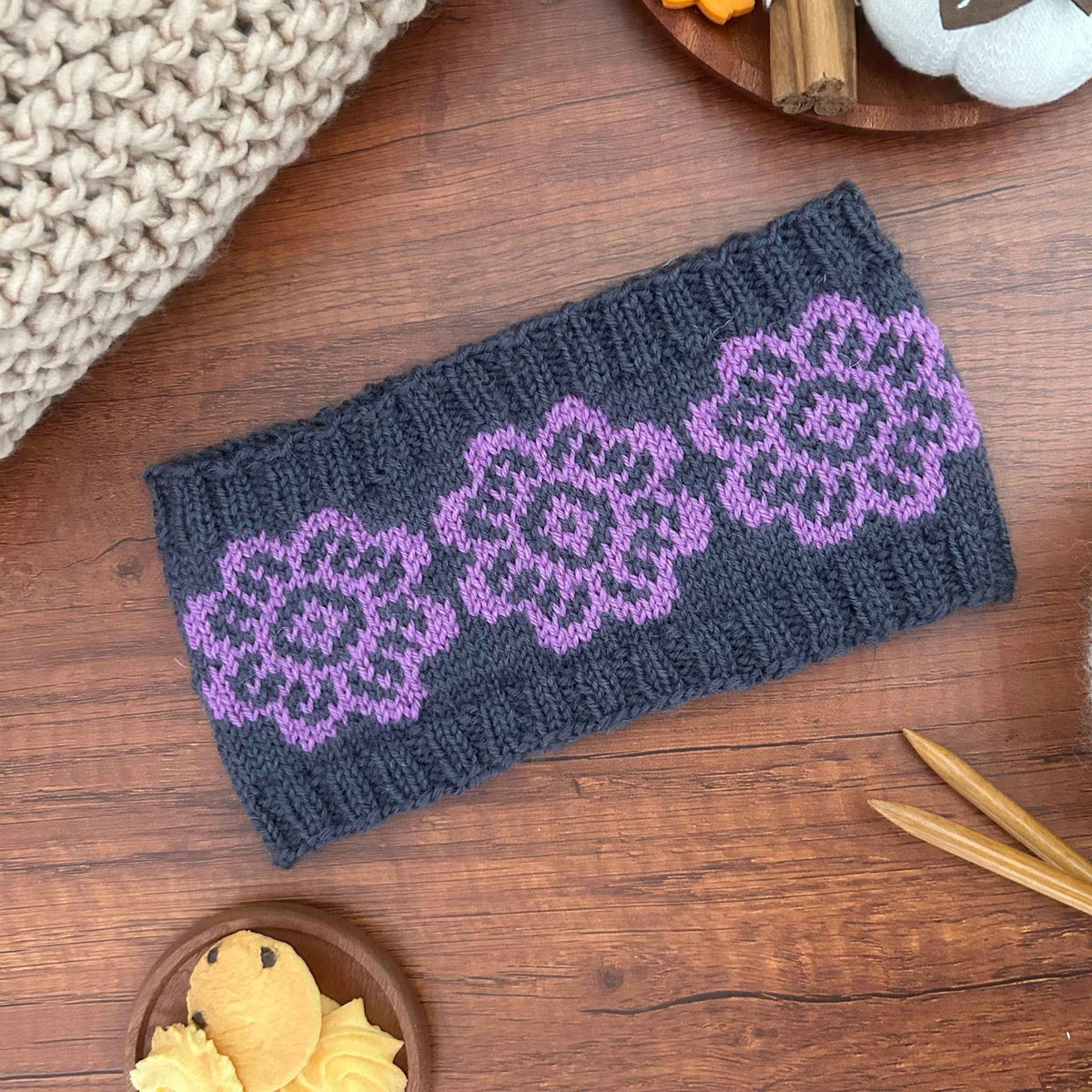 A dark charcoal gray knitted headband with three purple knit floral patterns, laid flat on a wooden table surrounded by knitting props.