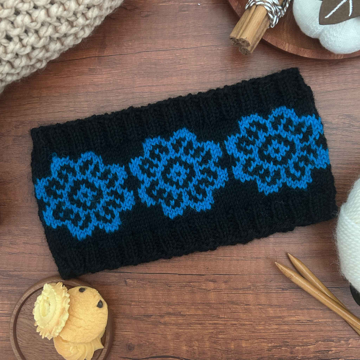 A black knit ear warmer or headband with three bright blue floral patterns, laid flat on a wooden surface with knitting props and yarn visible around it.