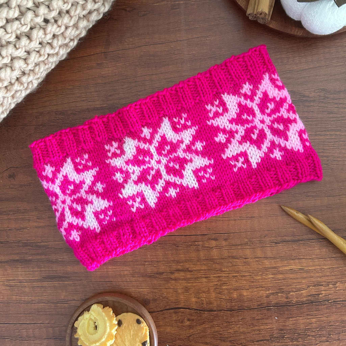 A bright pink knitted headband or ear warmer with a traditional white Selbu Star Norwegian snowflake pattern, laid flat on a dark wooden table with knitting props.