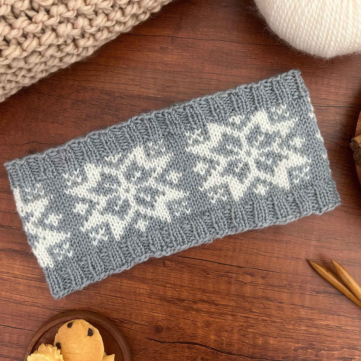 A rectangular gray knitted ear warmer or headband with a traditional white Norwegian Selbu star or snowflake pattern, laid flat on a wooden surface with knitting props around it