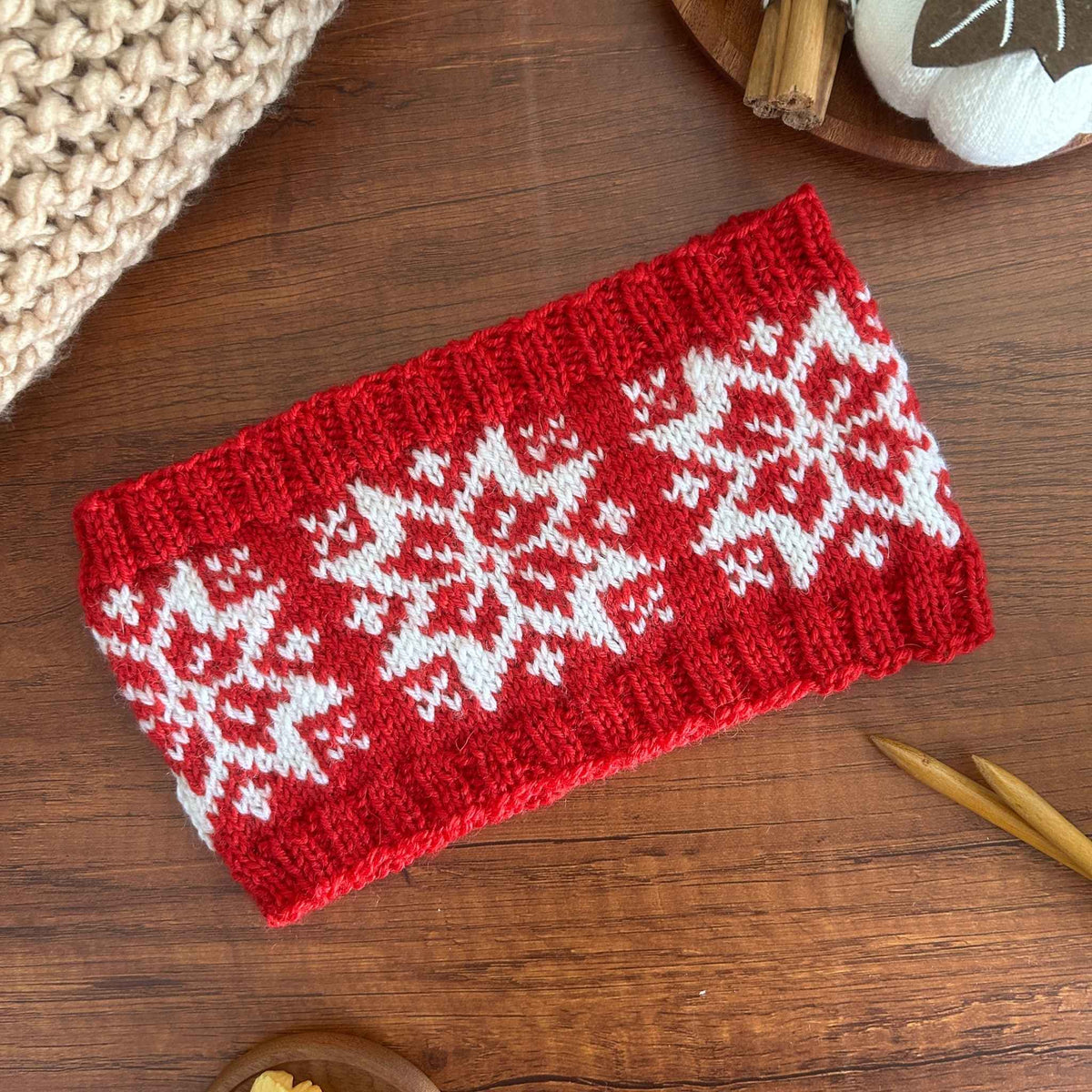 A rectangular red and white knitted ear warmer or headband with a traditional white Norwegian Selbu star or snowflake pattern, laid flat on a wooden surface with knitting props around it