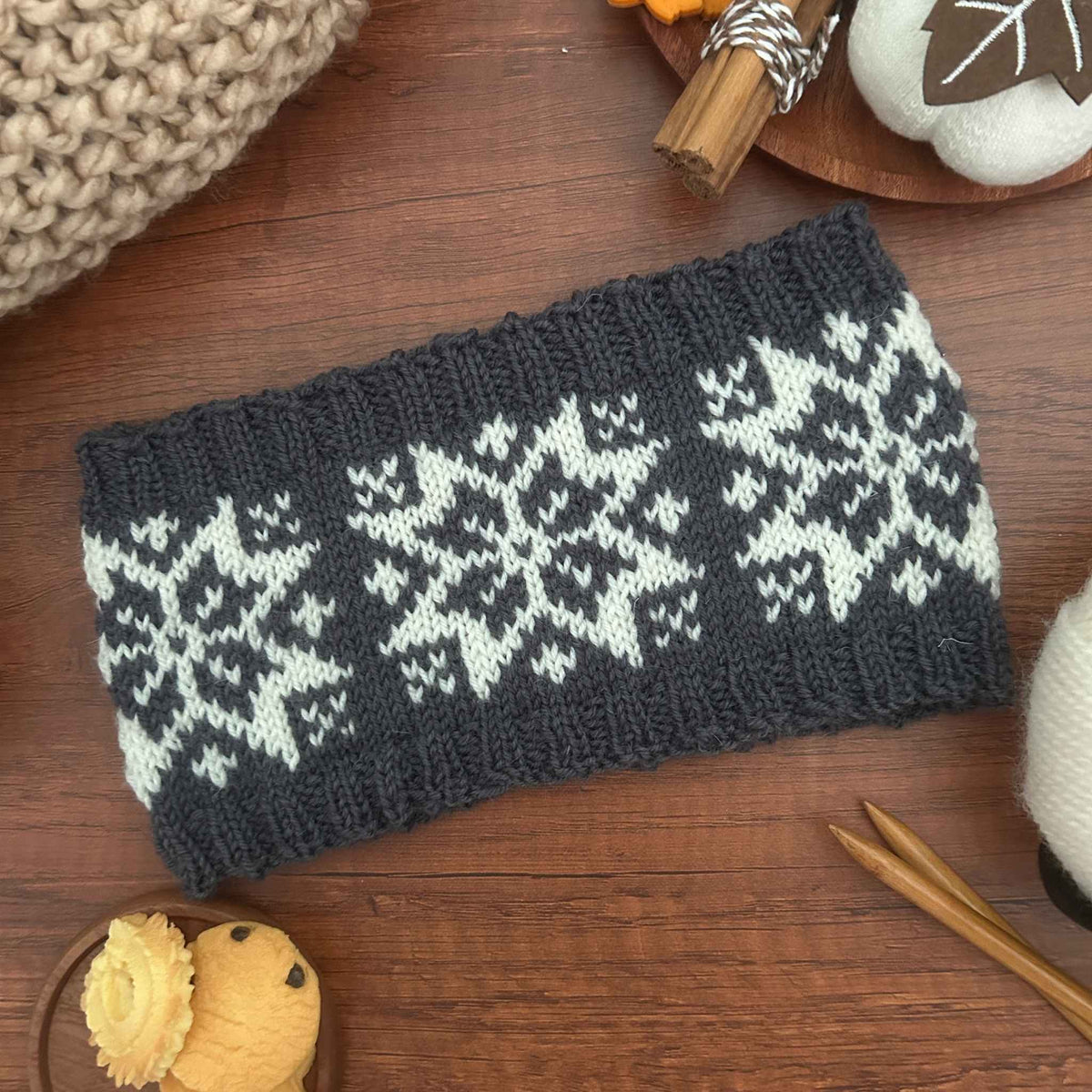 A rectangular gray knitted ear warmer or headband with a traditional white Norwegian Selbu star or snowflake pattern, laid flat on a wooden surface with knitting props around it