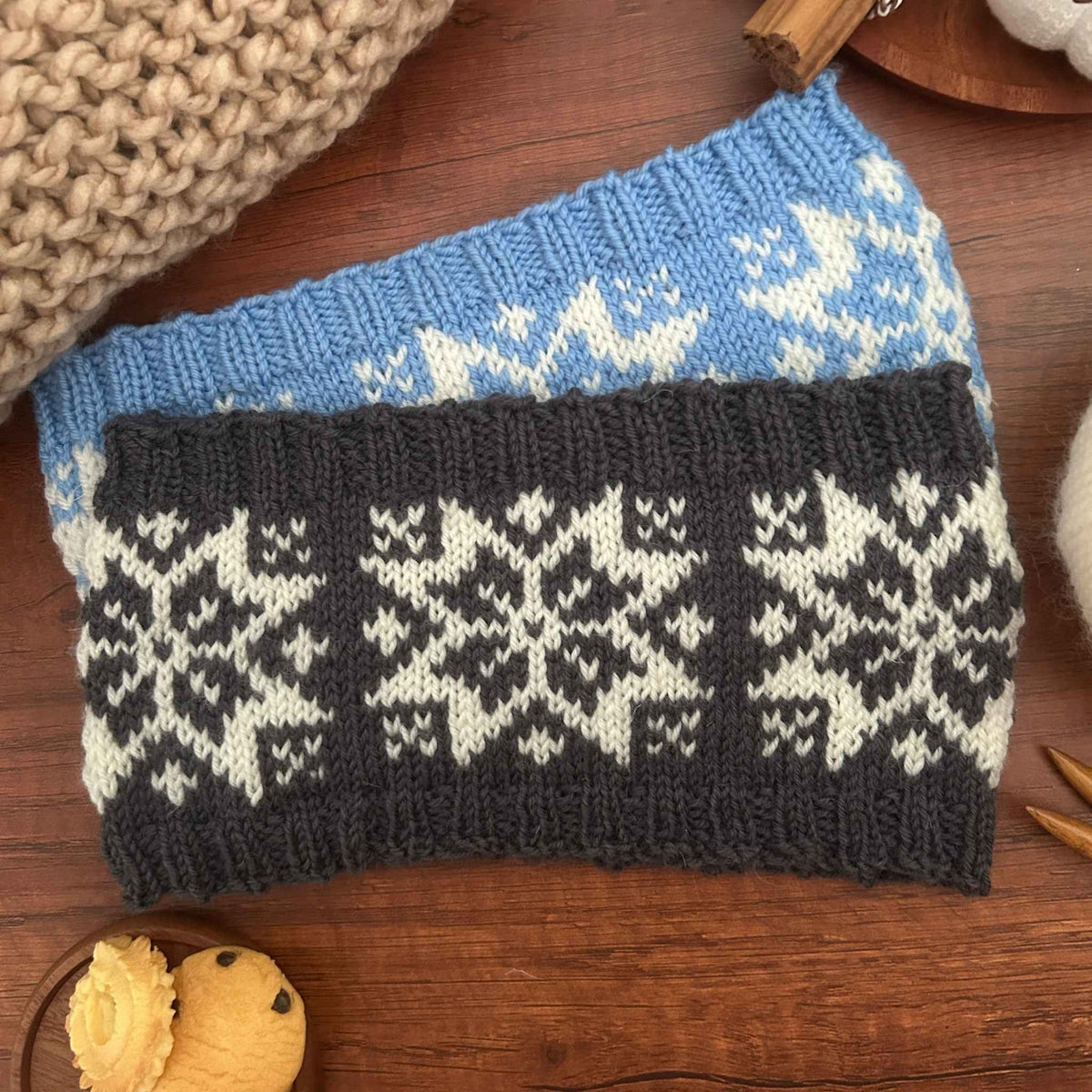 Two rectangular knitted ear warmers or headbands in dark grey and blue with a traditional white Norwegian Selbu star or snowflake pattern, laid flat on a wooden surface with knitting props around it