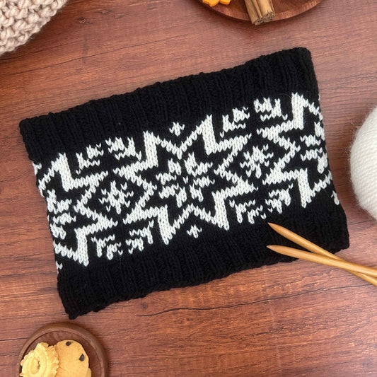 A black knit cowl or neck warmer with a traditional Norwegian stranded colourwork design of the Selbu Rose / Star, styled on a wooden table with knitting needles and yarn.