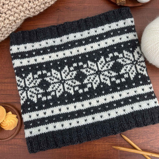 A square knitted scarf or cowl with a dark charcoal gray and off-white Nordic/Fair Isle pattern featuring the traditional Norwegian motif of a Selbu star or rose, and stripes, laid flat on a wooden surface with knitting props.