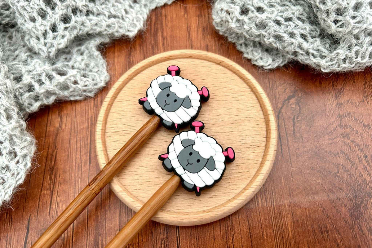 Two wooden knitting needles with sheep-shaped stoppers rest on a round wooden tray, set on a dark wood tabletop with a gray knitted blanket in the background.