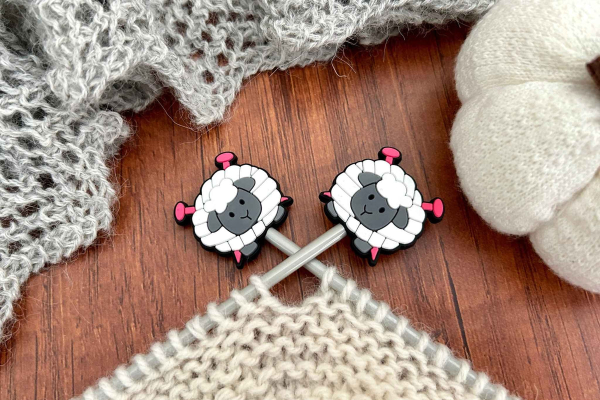 Two small sheep-shaped knitting needles stoppers resting on wooden knitting needles over a knitted cream piece, with gray knitted fabric and a white pumpkin in the background.