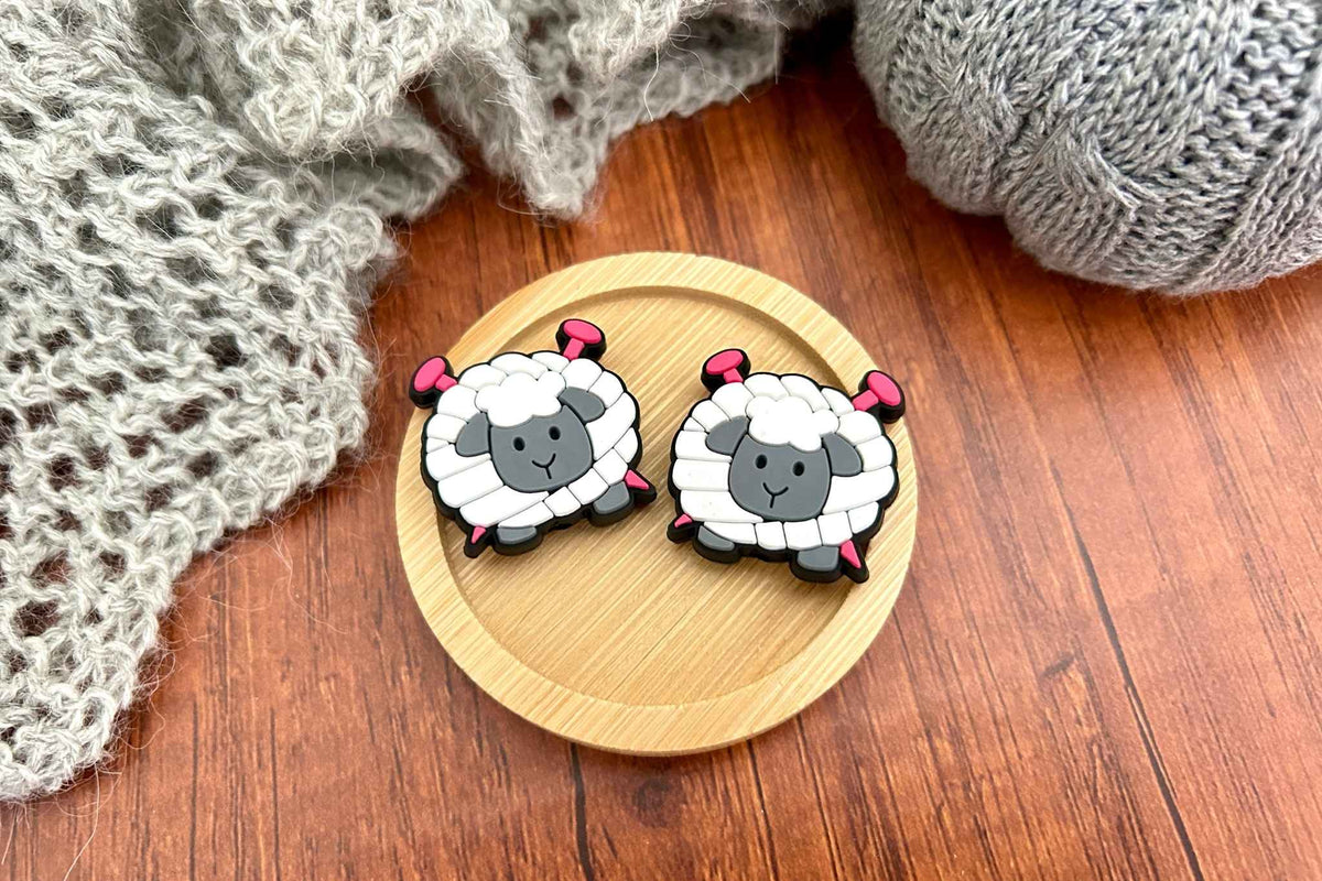 Two cute cartoon sheep stitch savers with gray faces, and pink cartoon knitting needles through them, displayed on a round wooden tray with a cozy gray knitted blanket and wooden surface in the background.