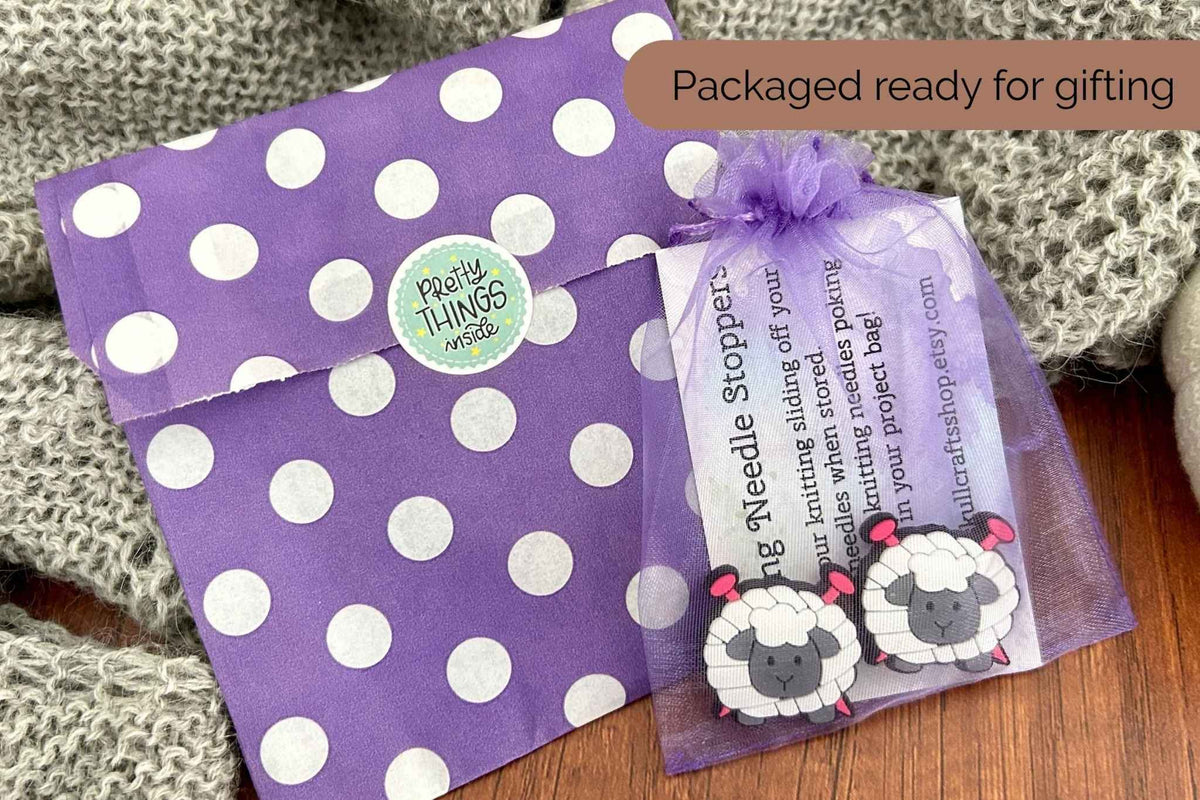 A purple and white polka dot gift bag with a round 'Pretty Things inside' sticker, placed next to a sheer purple gift bag holding two sheep-shaped needle stoppers, and an instruction card, on a wooden surface with a gray knit blanket.