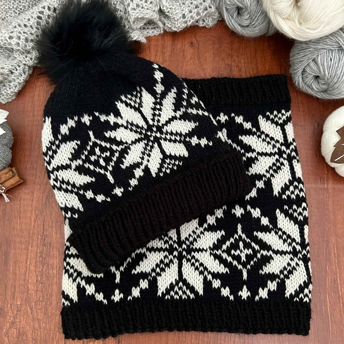 A black and cream knit beanie with a matching pom-pom and a matching knit scarf, laid flat on a wooden surface with yarn balls and a knitted blanket in the background. The motif is a traditional Norwegian Selbu star, often found in stranded colourwork designs