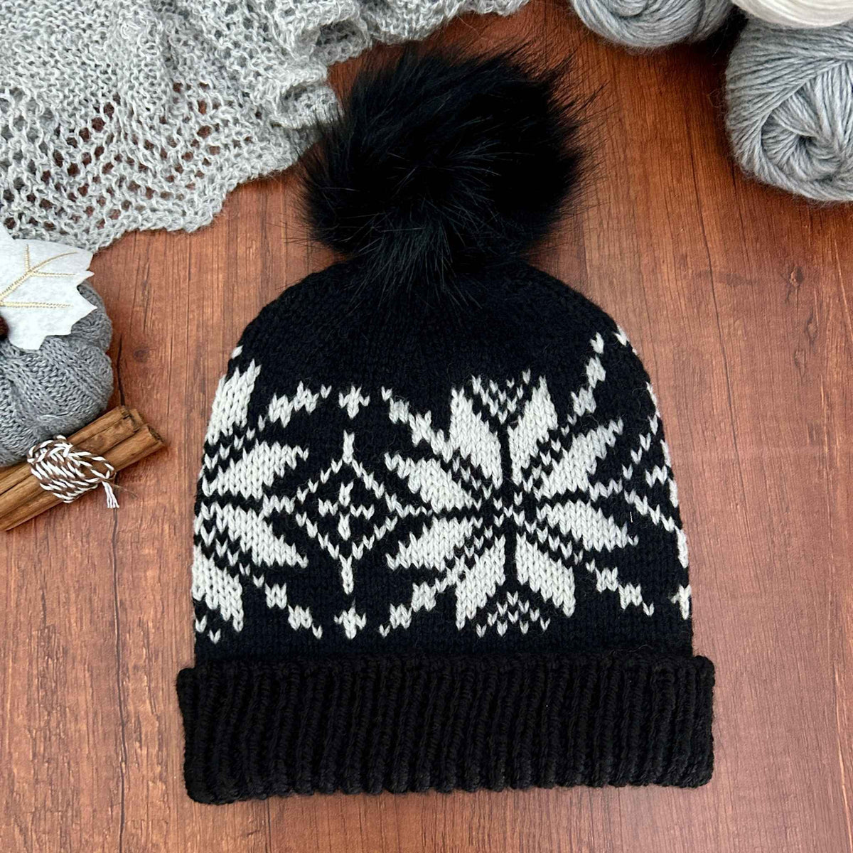 A black knit beanie with a white Selbu rose. star, or snowflake pattern and a fluffy black pom-pom, laid flat on a wooden surface with yarn and a knitted scarf in the background. The motif is a traditional Norwegian stranded colourwork design