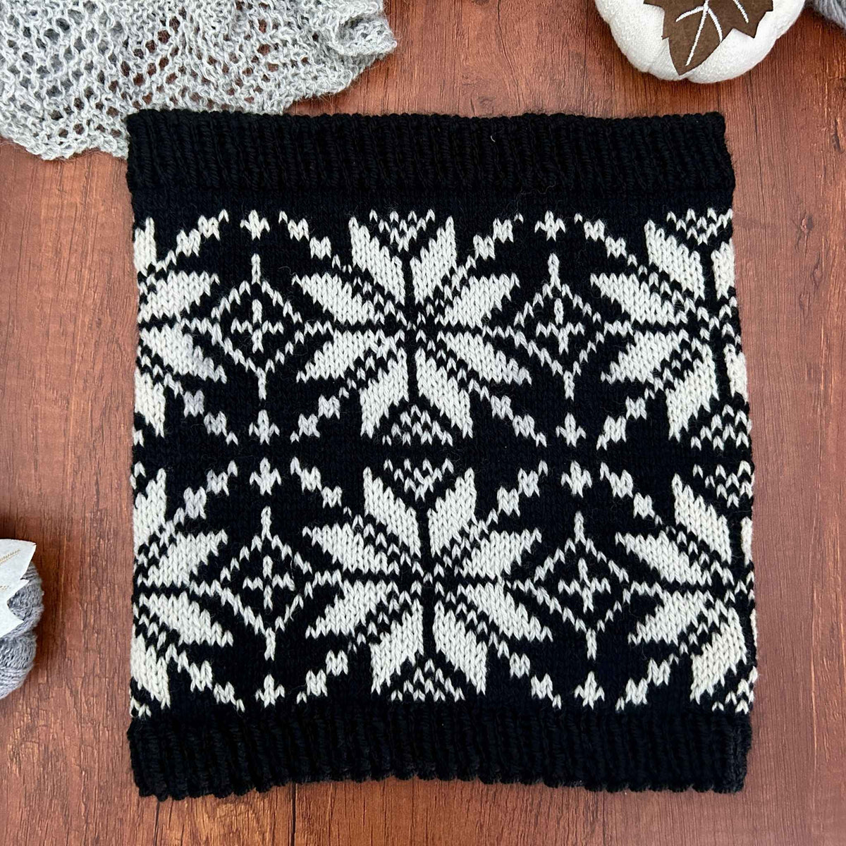 A square black and white knit cowl with a traditional Nordic star or snowflake pattern, laid flat on a wooden surface with yarn and a crocheted piece as props. Knitted in stranded colorwork