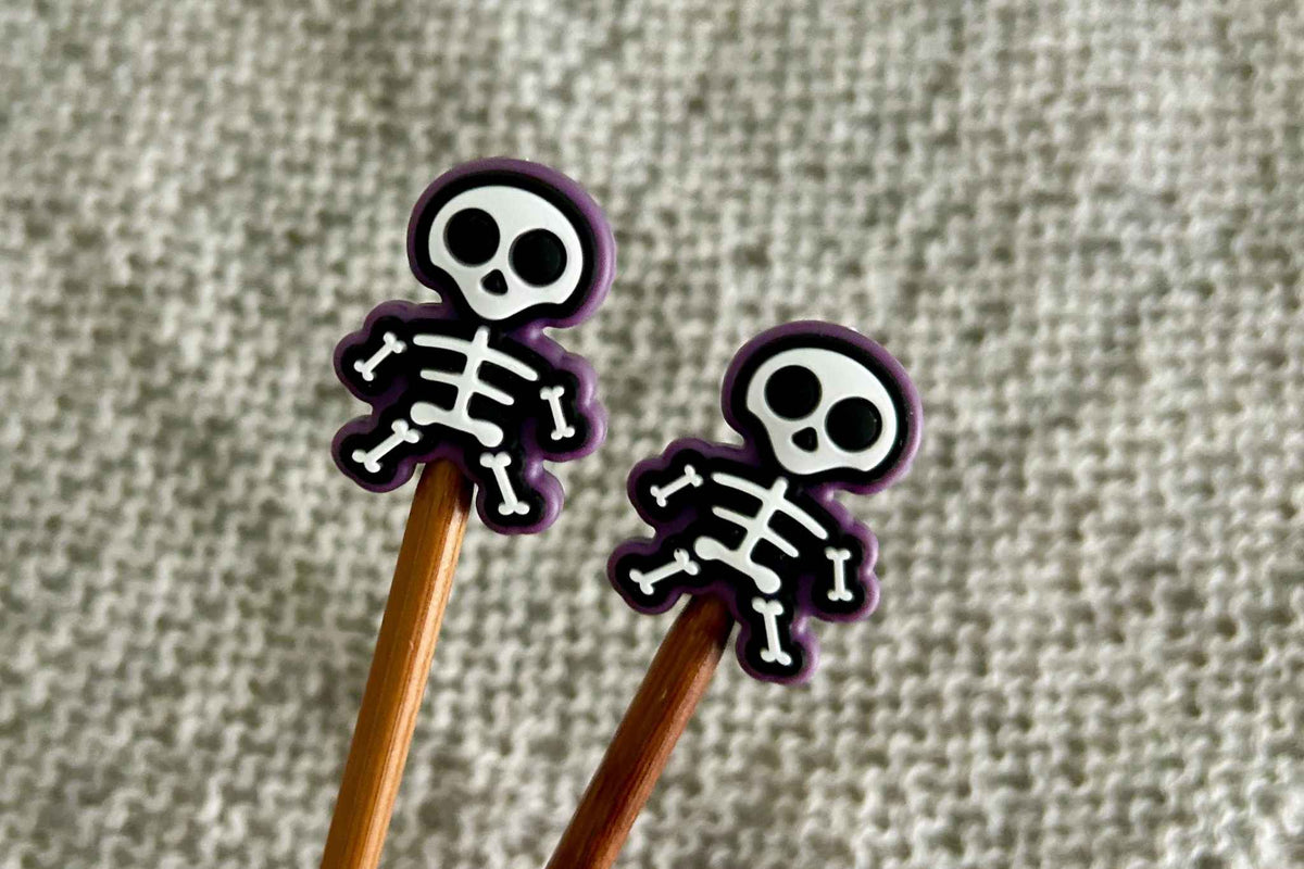 Two wooden knitting needles with skeleton knitting needle stoppers on the tips, photographed against a textured grey fabric background.