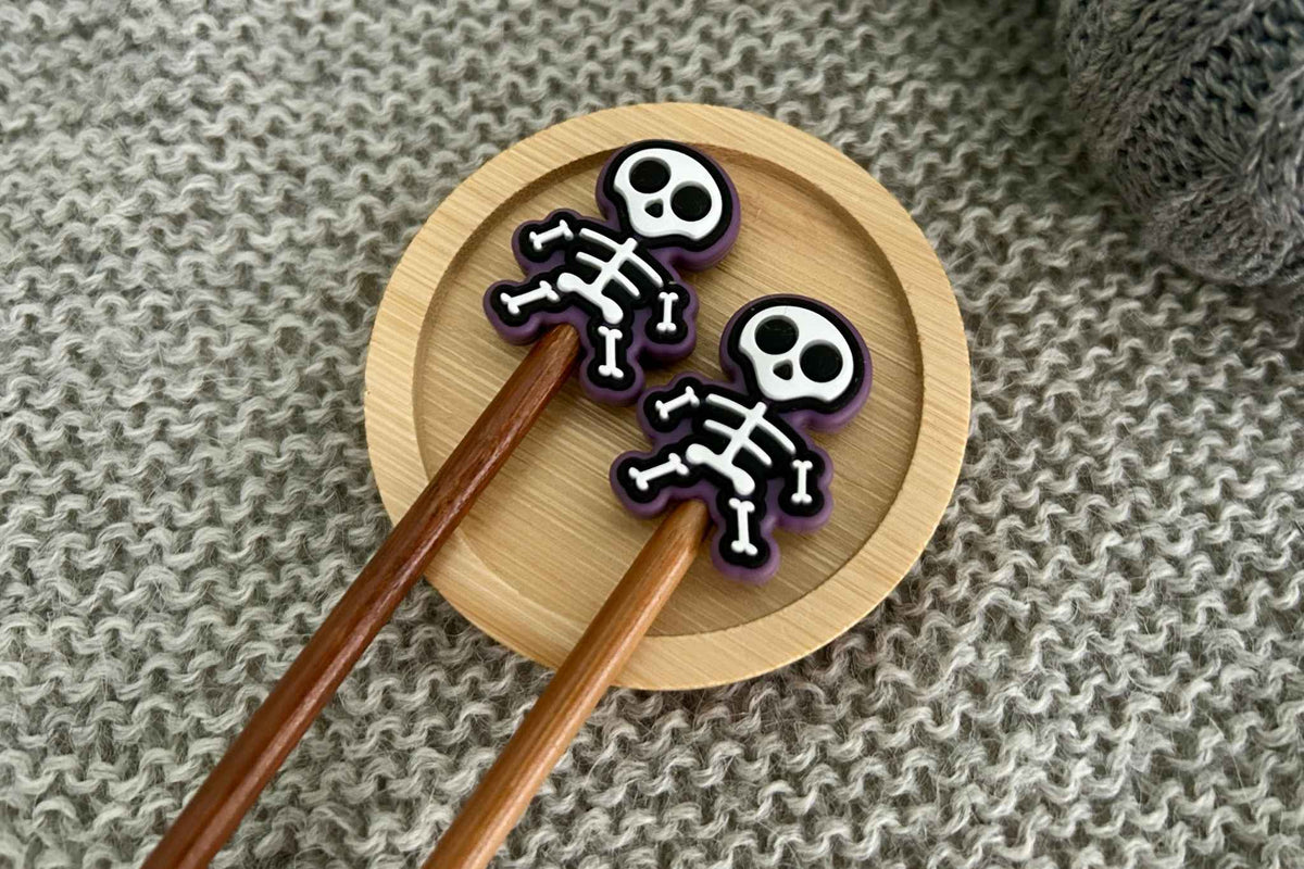 Two small skeleton-shaped knitting needle stoppers with purple trim and white bone details rest on a round wooden tray, with brown knitting needles extending downward on a gray knitted fabric background.