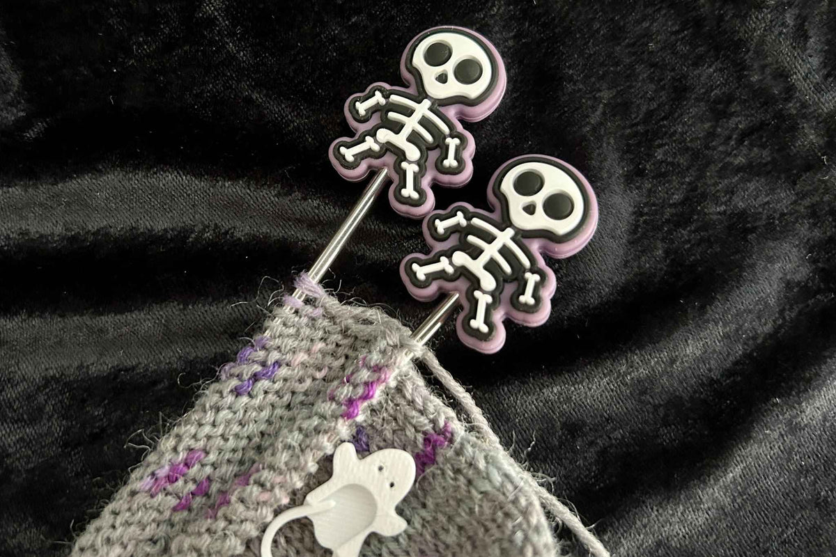Two metal knitting needles with skull-shaped tips and a gray knitted fabric with purple yarn accents, set against a dark textured background.