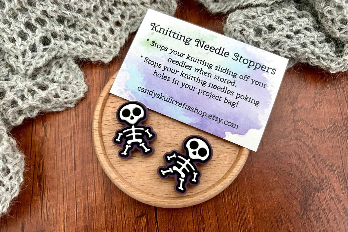 Two small black-and-white skeleton-shaped knitting needle stoppers displayed on a round wooden coaster next to a printed product card, with a knitted gray textile draped in the background on a wooden surface.