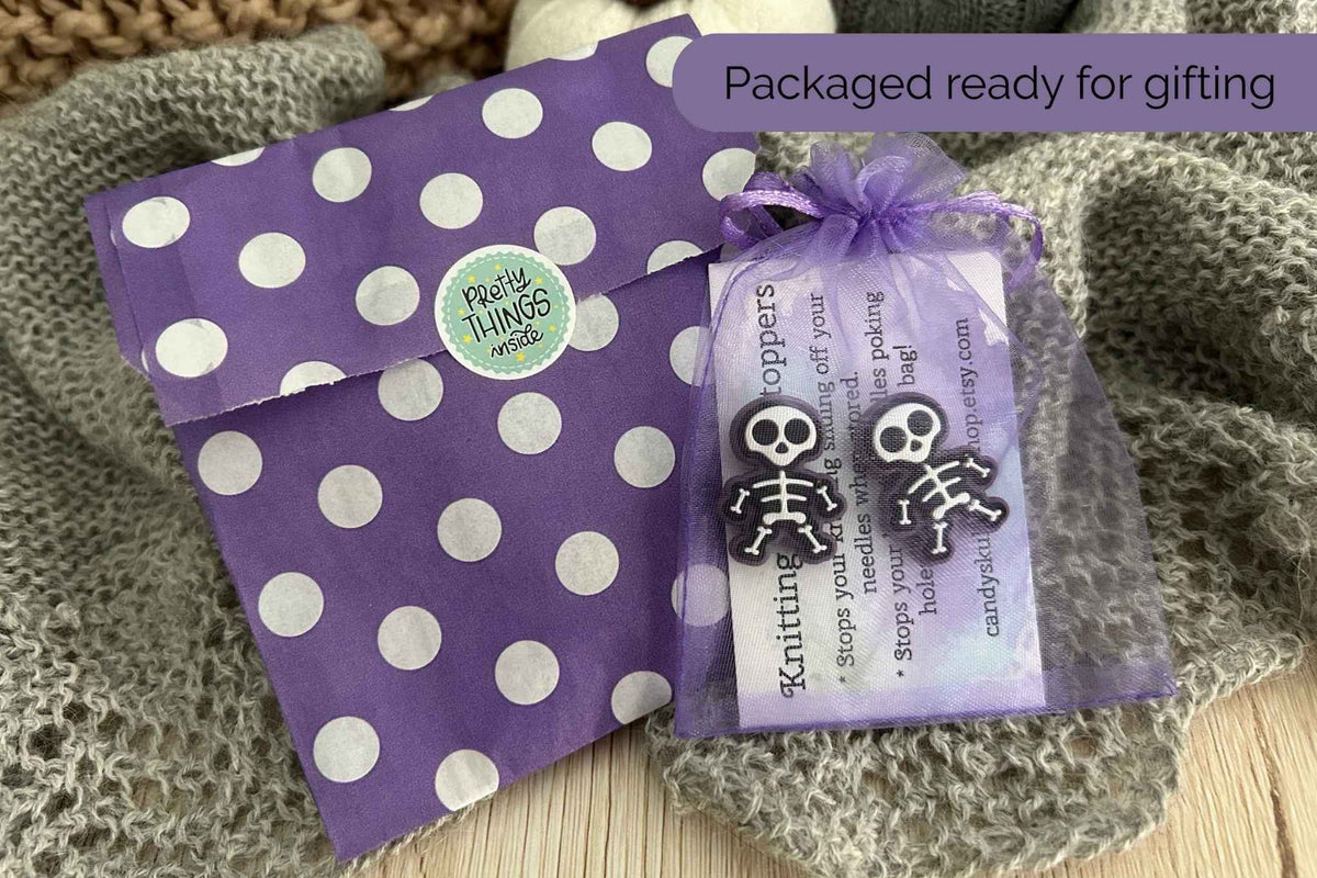 Purple and white polka dot gift packaging with a small purple organza bag tied with a ribbon, featuring a black-and-white skeleton knitting needle stoppers, an instruction card, and a title that reads 'Packaged ready for gifting.' The items are styled on a knitted gray blanket.